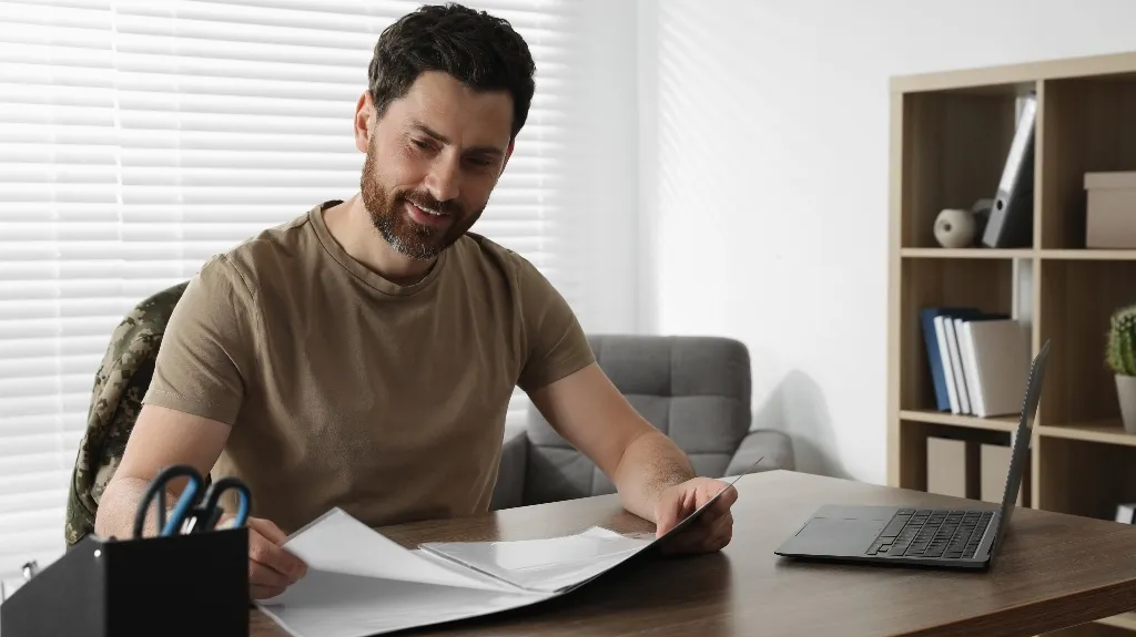 Man in casual attire reviewing documents at a wooden desk with laptop and office supplies.