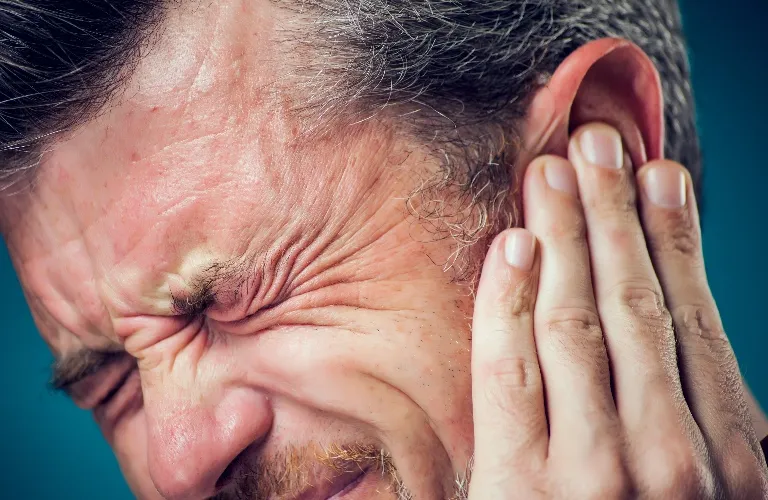 Close-up of a man with gray hair pressing his hand against his ear in discomfort.