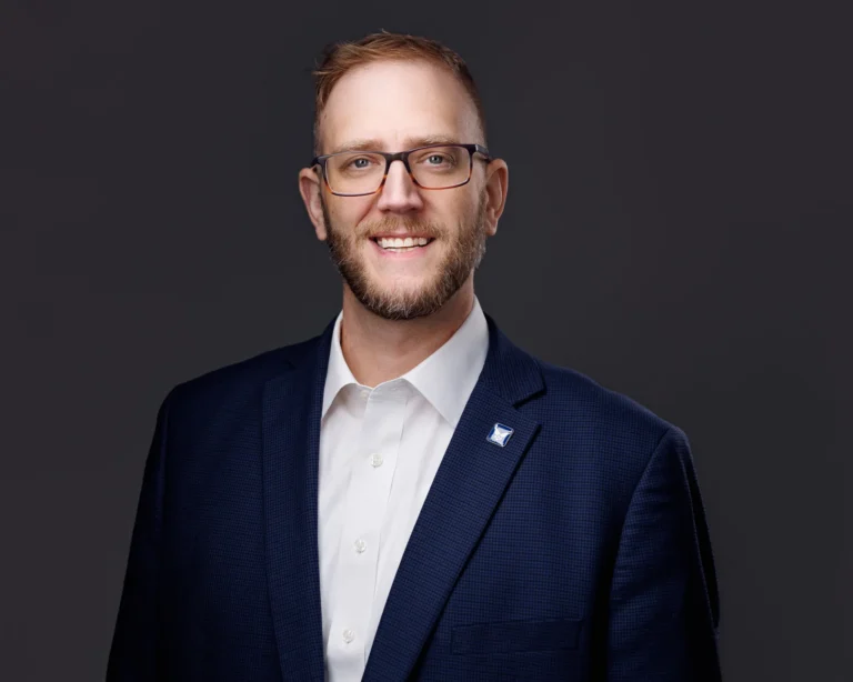 Professional headshot of a smiling man with glasses, beard, navy blazer, and white shirt.