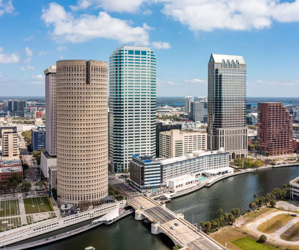 Aerial view of a city skyline with tall modern buildings, a river, and a bridge under blue sky.
