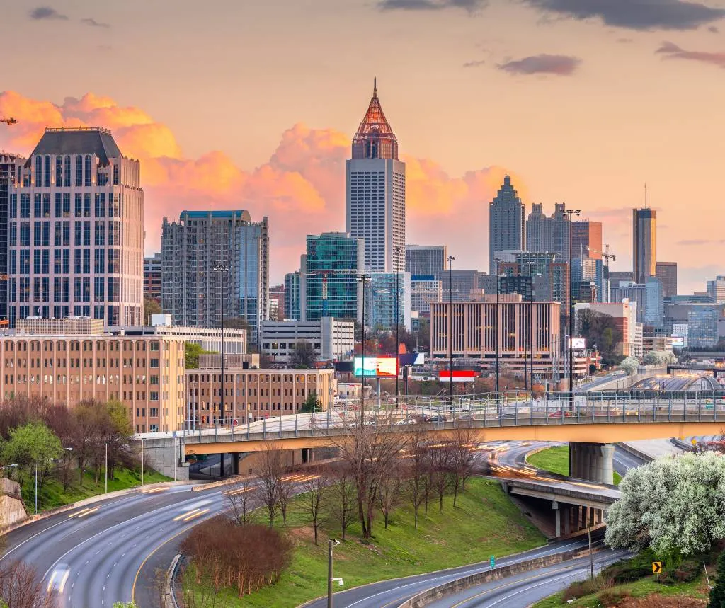 City skyline at sunset featuring modern high-rise buildings and a curving highway with light traffic.