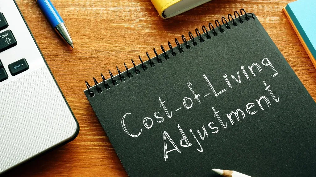 Black spiral notebook with white text "Cost-of-Living Adjustment" on wooden desk, beside laptop and pen.