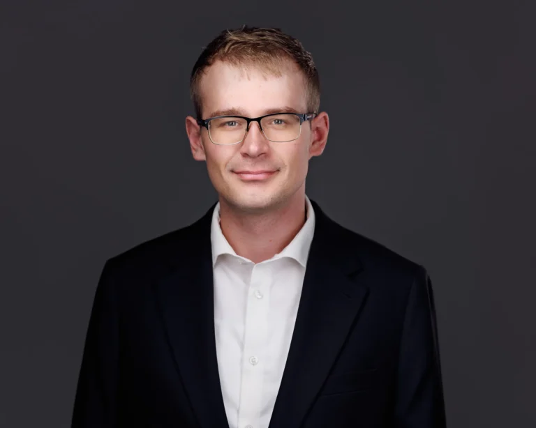 Professional headshot of young man with short light brown hair, glasses, white shirt, and dark blazer.