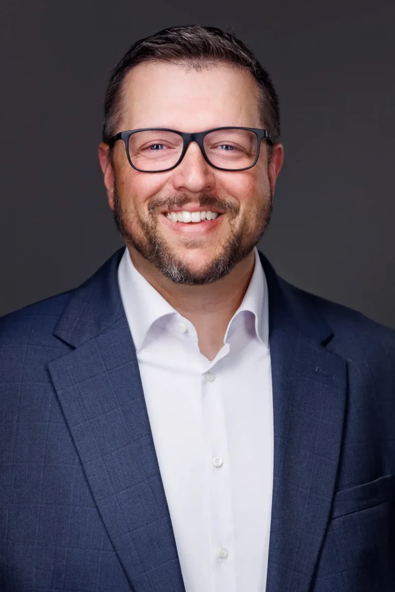 Professional headshot of a smiling man with glasses, beard, white shirt, and navy blue suit.