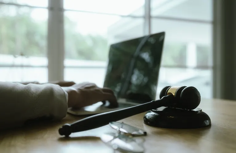 Wooden judge's gavel on desk with blurred laptop and hand in background.