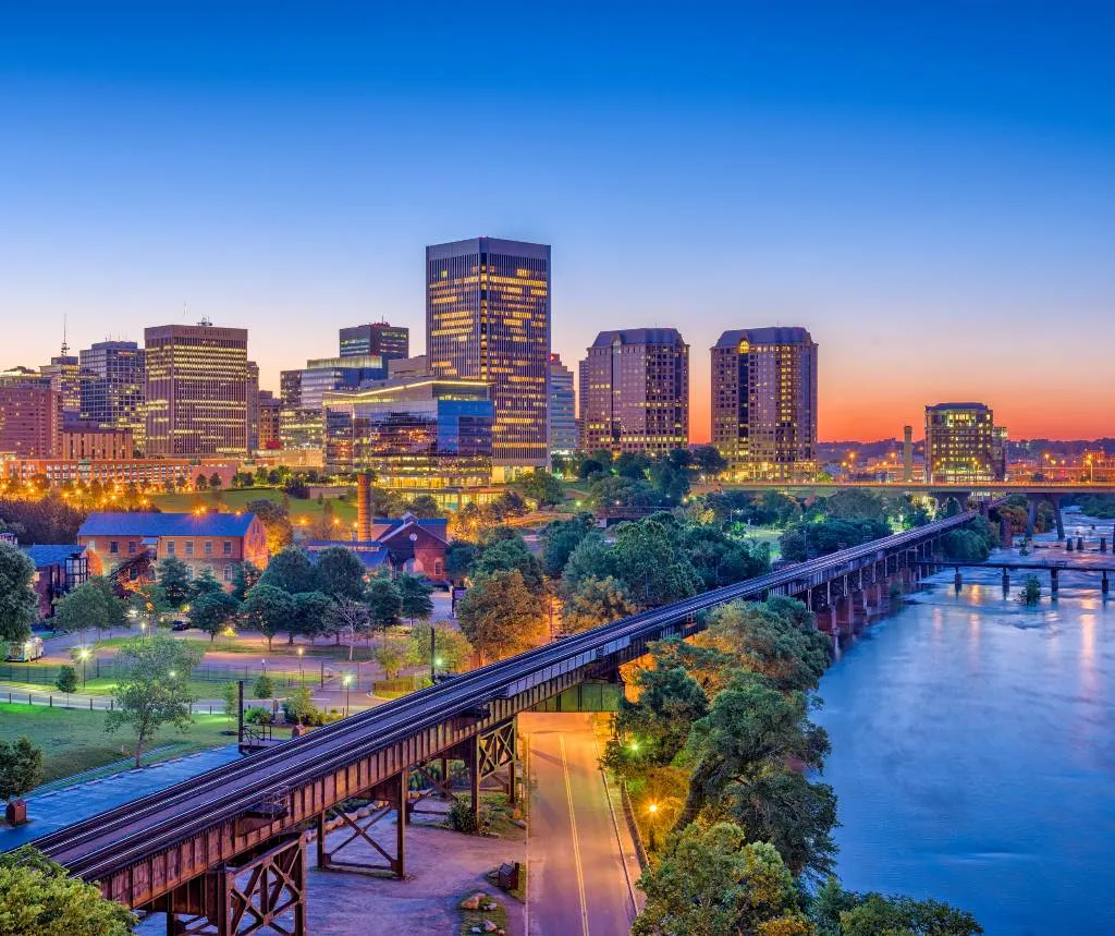 City skyline at dusk with illuminated buildings, a river, and a railway bridge.