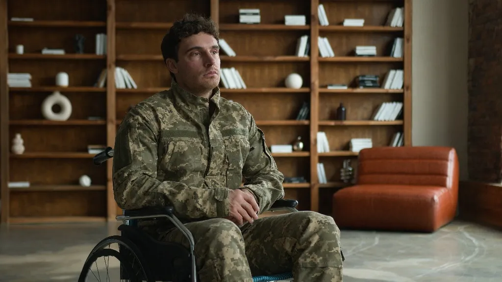 Man in camouflage military uniform seated in wheelchair in modern room with wooden bookshelves and orange chair.