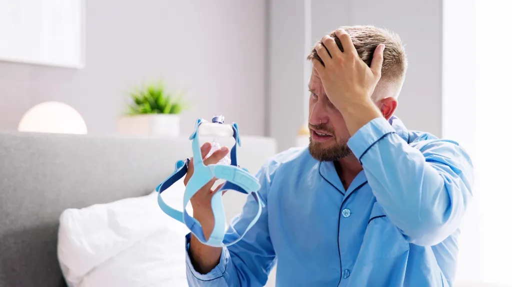 Man in blue pajamas sitting on bed, holding and examining a CPAP mask, appearing concerned.
