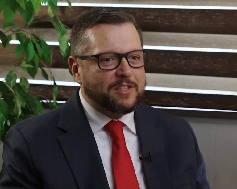 Man with glasses, beard, dark suit, red tie, smiling indoors near window blinds and green plant.