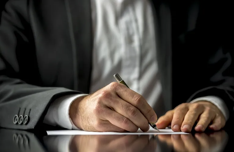 Close-up of a man in a suit writing on white paper with a pen at a reflective desk.
