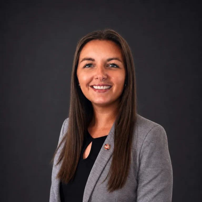 Professional headshot of a smiling woman with long brown hair wearing a gray blazer and black top.