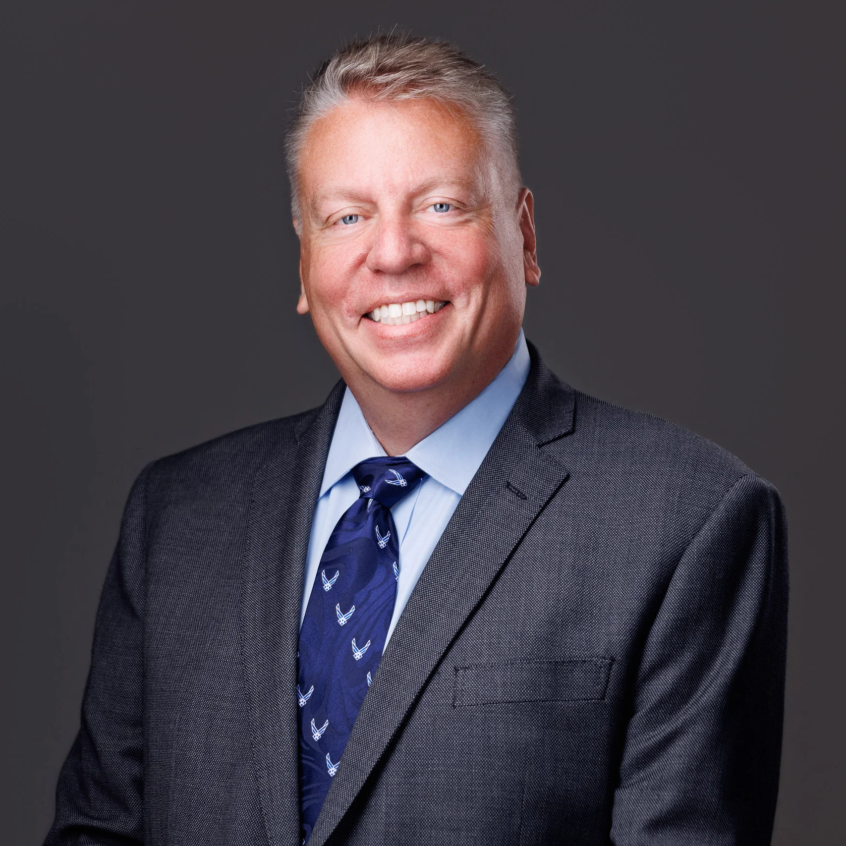 Professional portrait of a middle-aged man in a dark suit, blue shirt, and patterned tie against gray background.