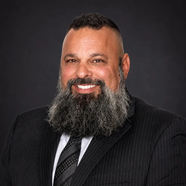 Professional portrait of a smiling man with a thick gray beard wearing a pinstripe suit and patterned tie.