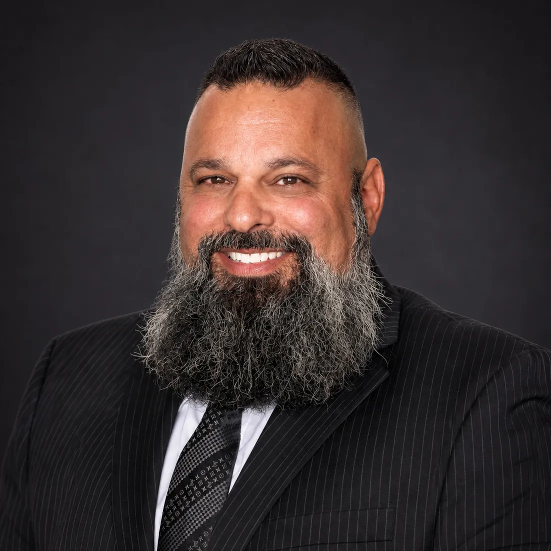 Professional portrait of a smiling man with a thick gray beard wearing a pinstripe suit and patterned tie.