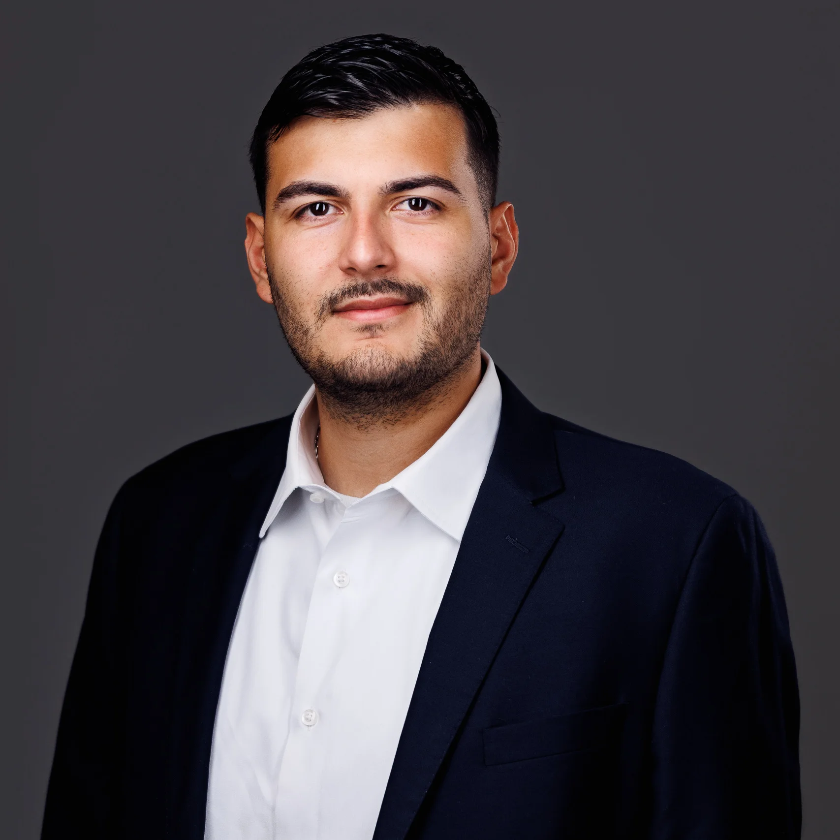 Professional headshot of a man with dark hair, beard, white shirt, and navy blazer on gray background.