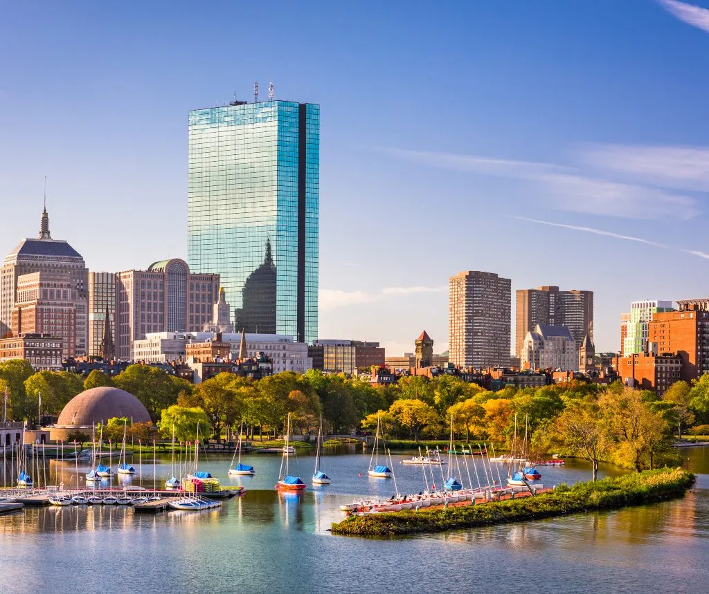 City skyline with modern skyscrapers and sailboats docked on a calm river under a clear blue sky.