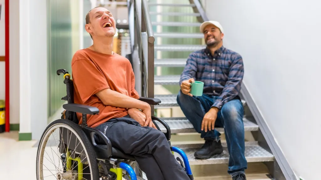 Two men smiling and laughing; one in a wheelchair wearing an orange shirt, the other sitting on stairs holding a green mug.