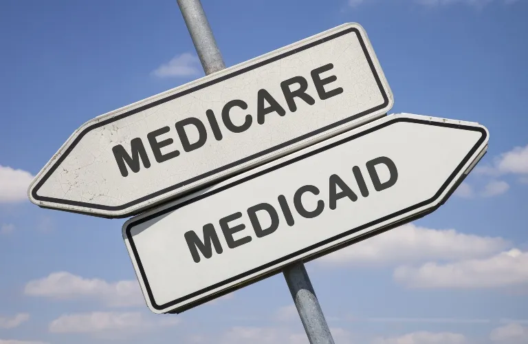 White directional road signs with black text reading "Medicare" and "Medicaid" against a blue sky background.