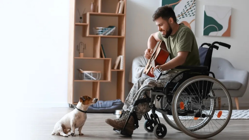Man in wheelchair playing guitar while small dog attentively watches indoors.