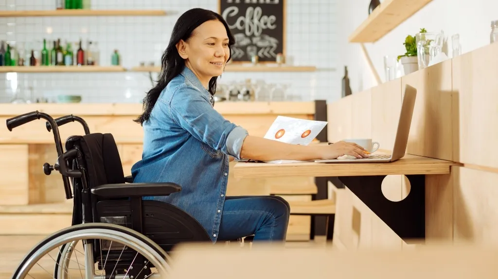 Woman in a wheelchair working on a laptop at a wooden café counter with coffee menu.