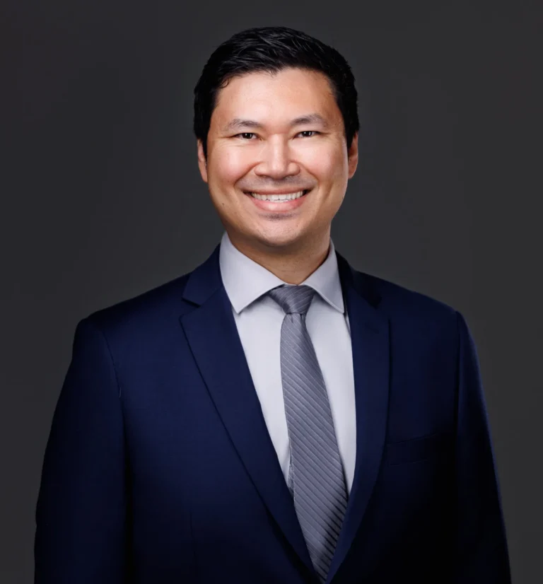 Professional headshot of a smiling man wearing a navy suit, light gray shirt, and gray tie.