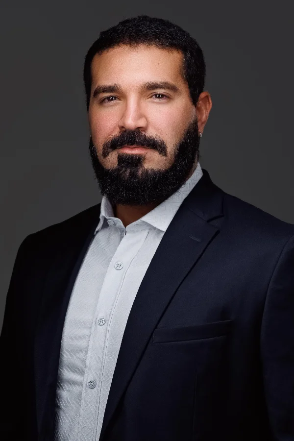 Professional headshot of a bearded man in a dark suit jacket and light dress shirt against a gray background.