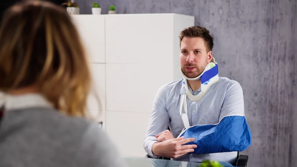 Man with arm in blue sling and neck brace sitting across from woman in office.