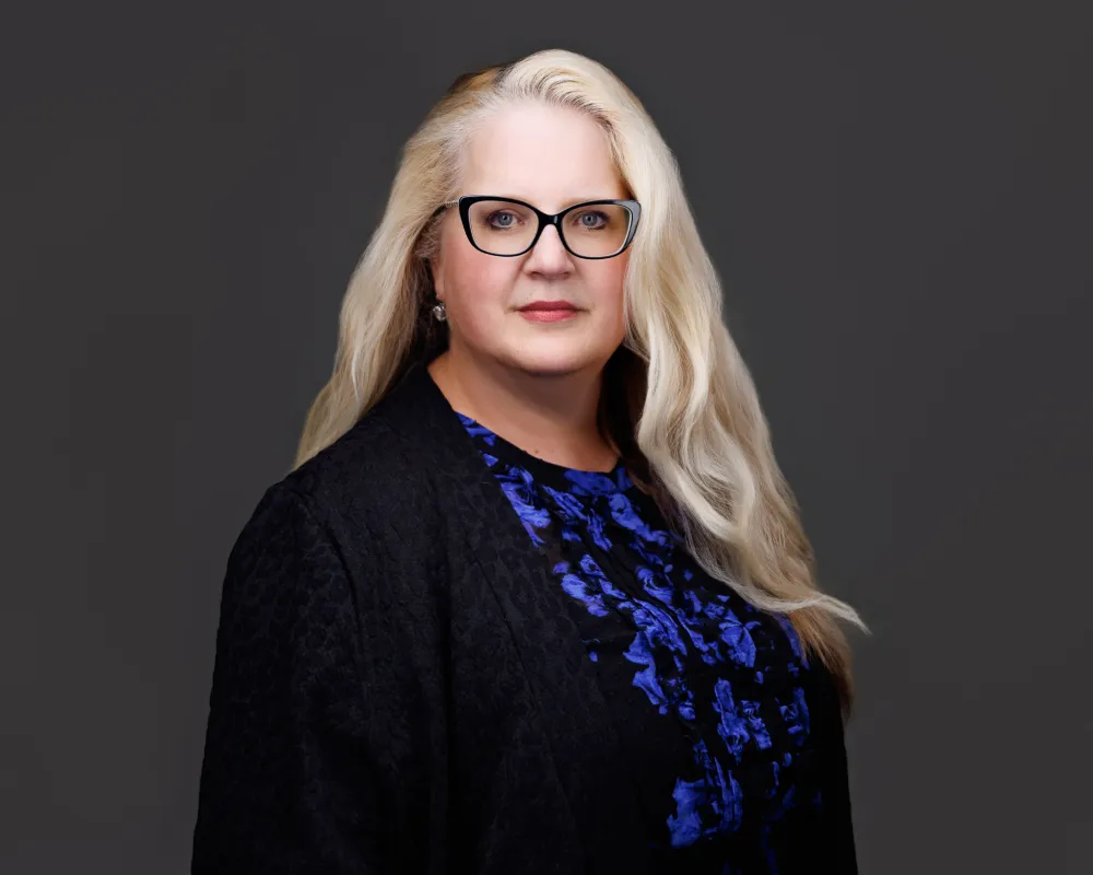 Professional portrait of a woman with long blonde hair, black glasses, and a blue patterned blouse.