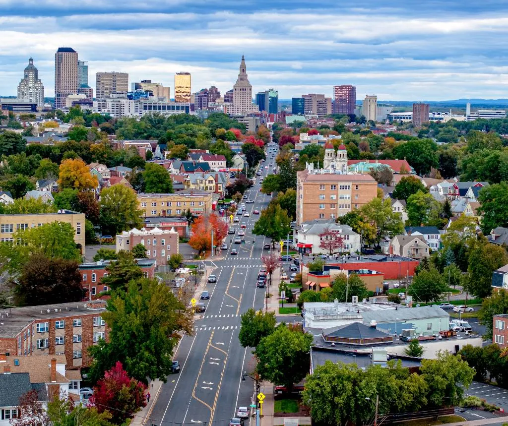 Aerial view of a cityscape with a central road, mixed residential and commercial buildings, and autumn foliage.