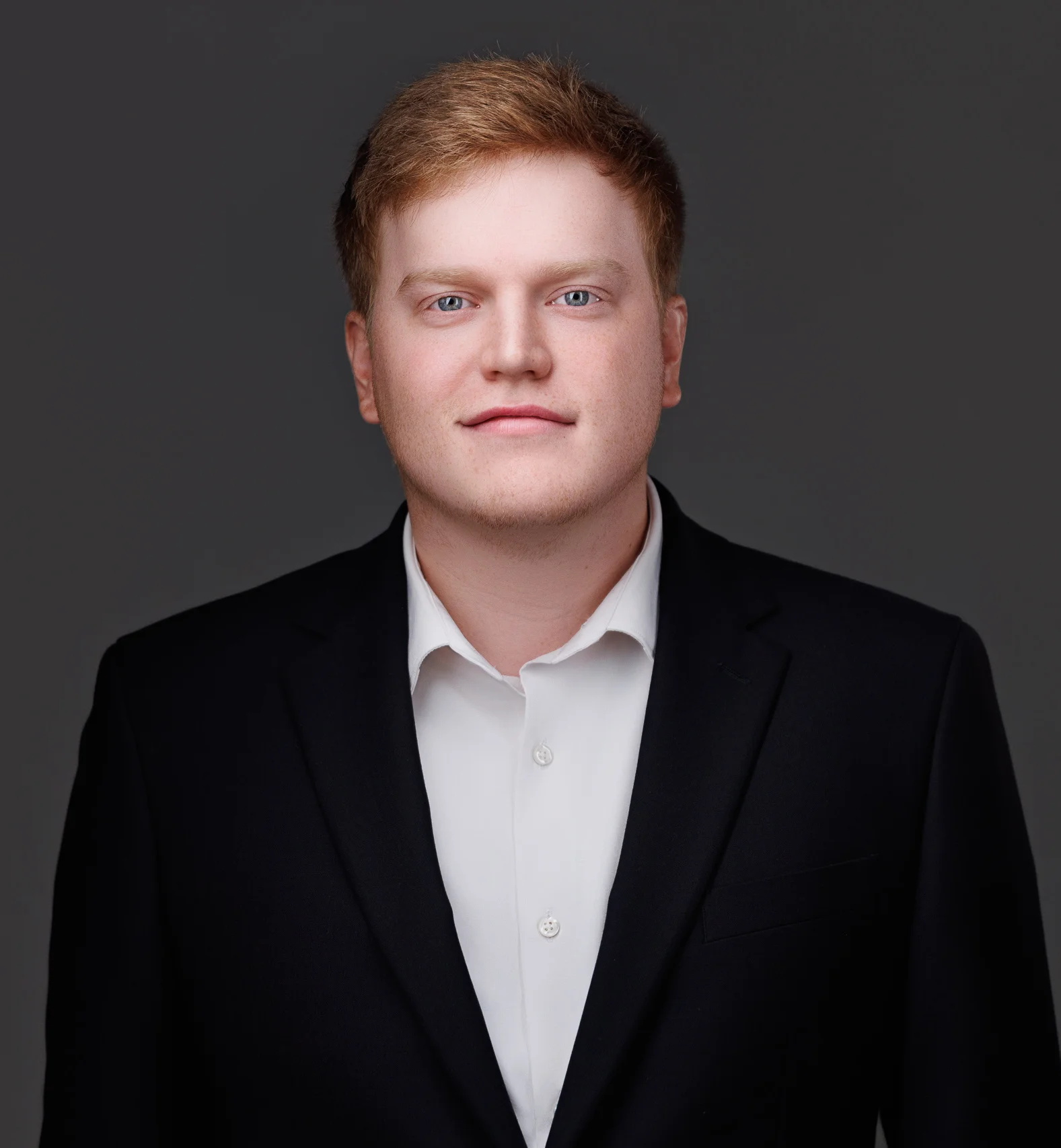 Professional portrait of a young man with red hair, blue eyes, wearing a black suit and white shirt.