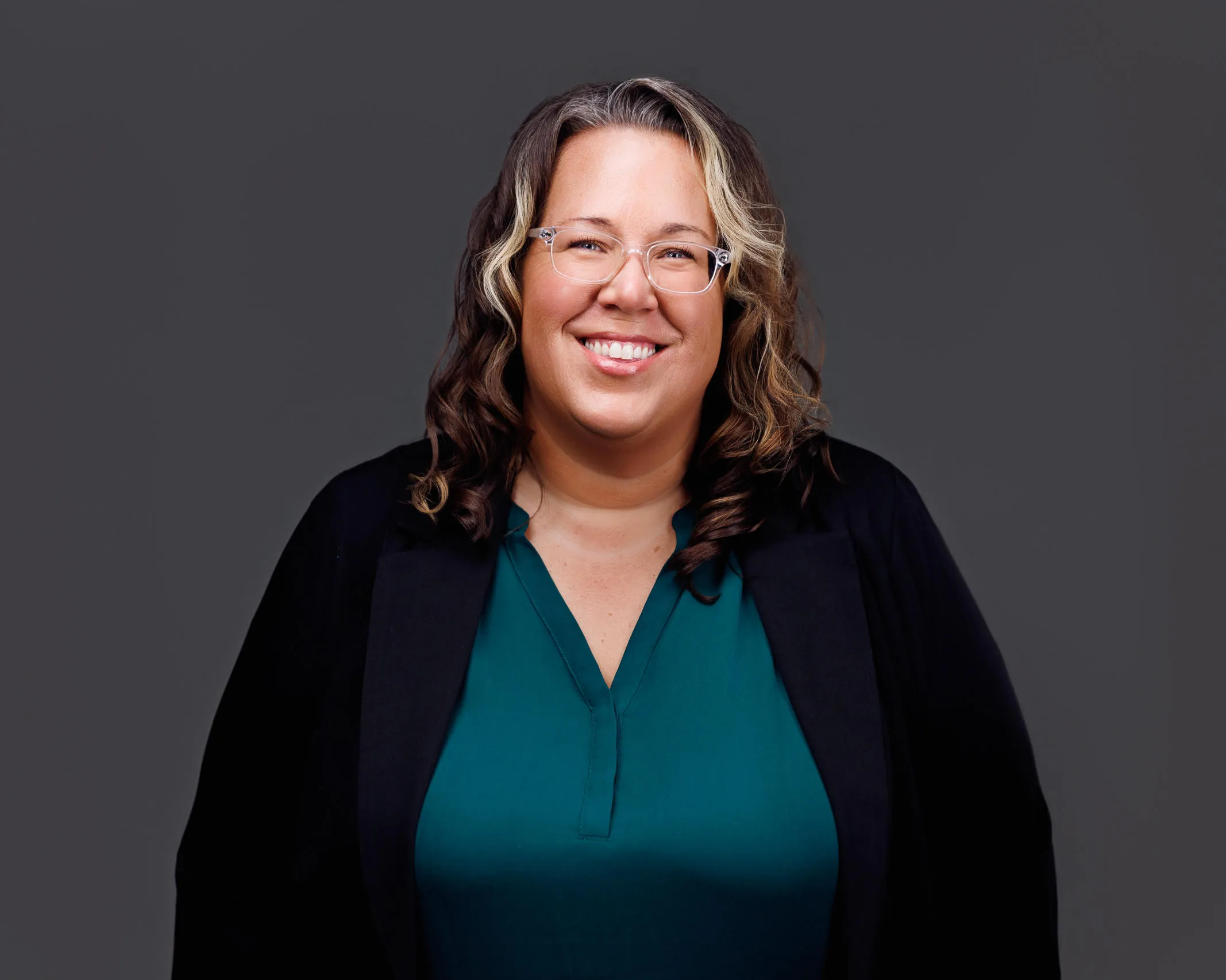 Professional headshot of a smiling woman with curly hair, glasses, teal blouse, and black blazer.
