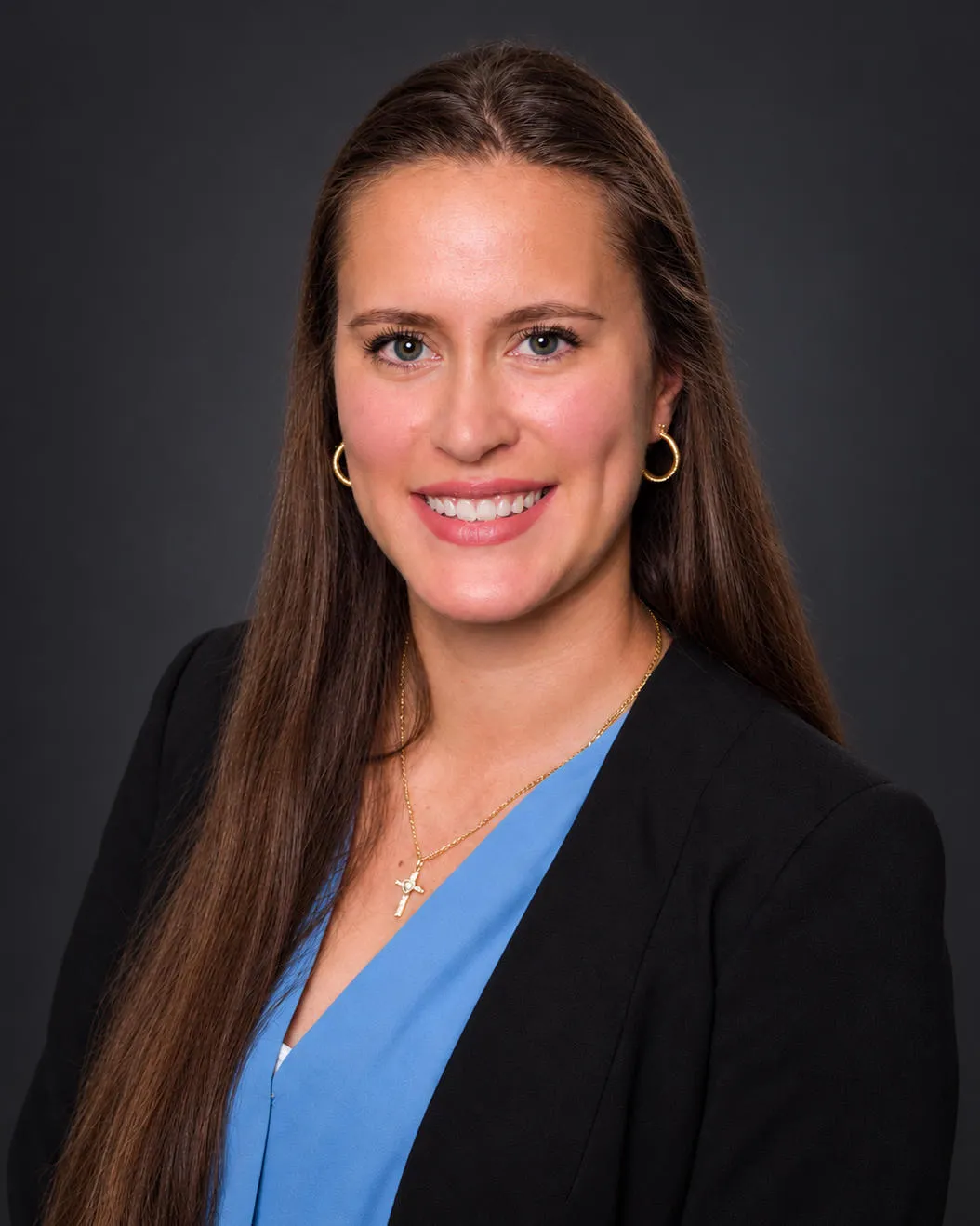 Professional headshot of a woman with long brown hair, wearing a black blazer, blue blouse, and gold accessories.