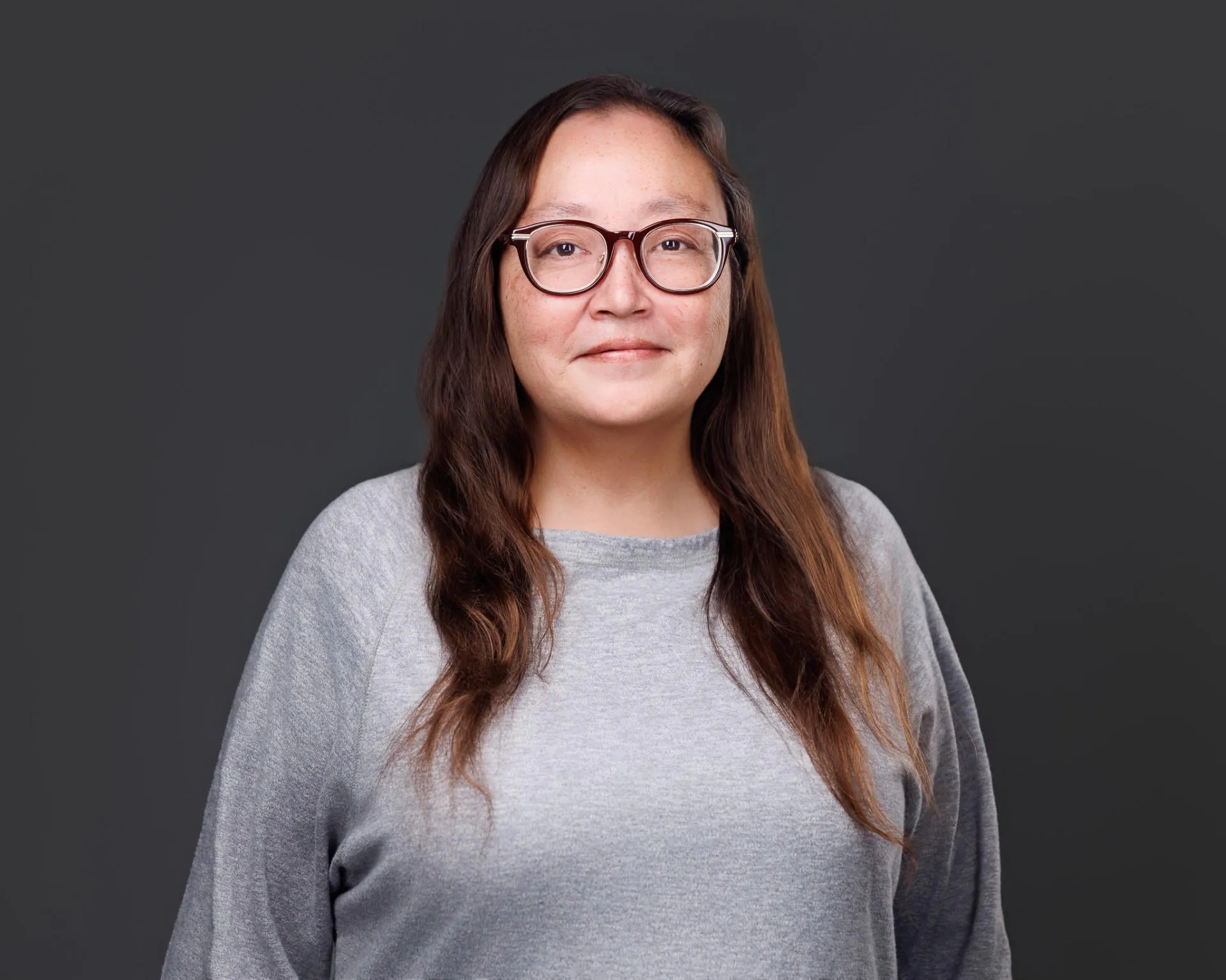Portrait of a woman with long brown hair, glasses, and a gray sweater against dark background.