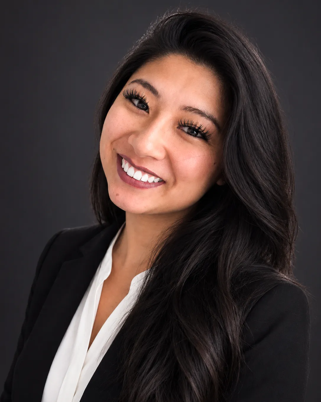 Professional headshot of a smiling woman with long dark hair, wearing a black blazer and white blouse.